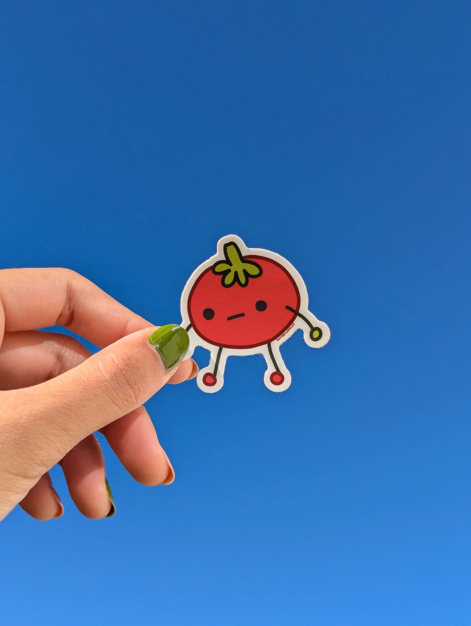 A hand holding up a cartoon tomato sticker against a clear sky background