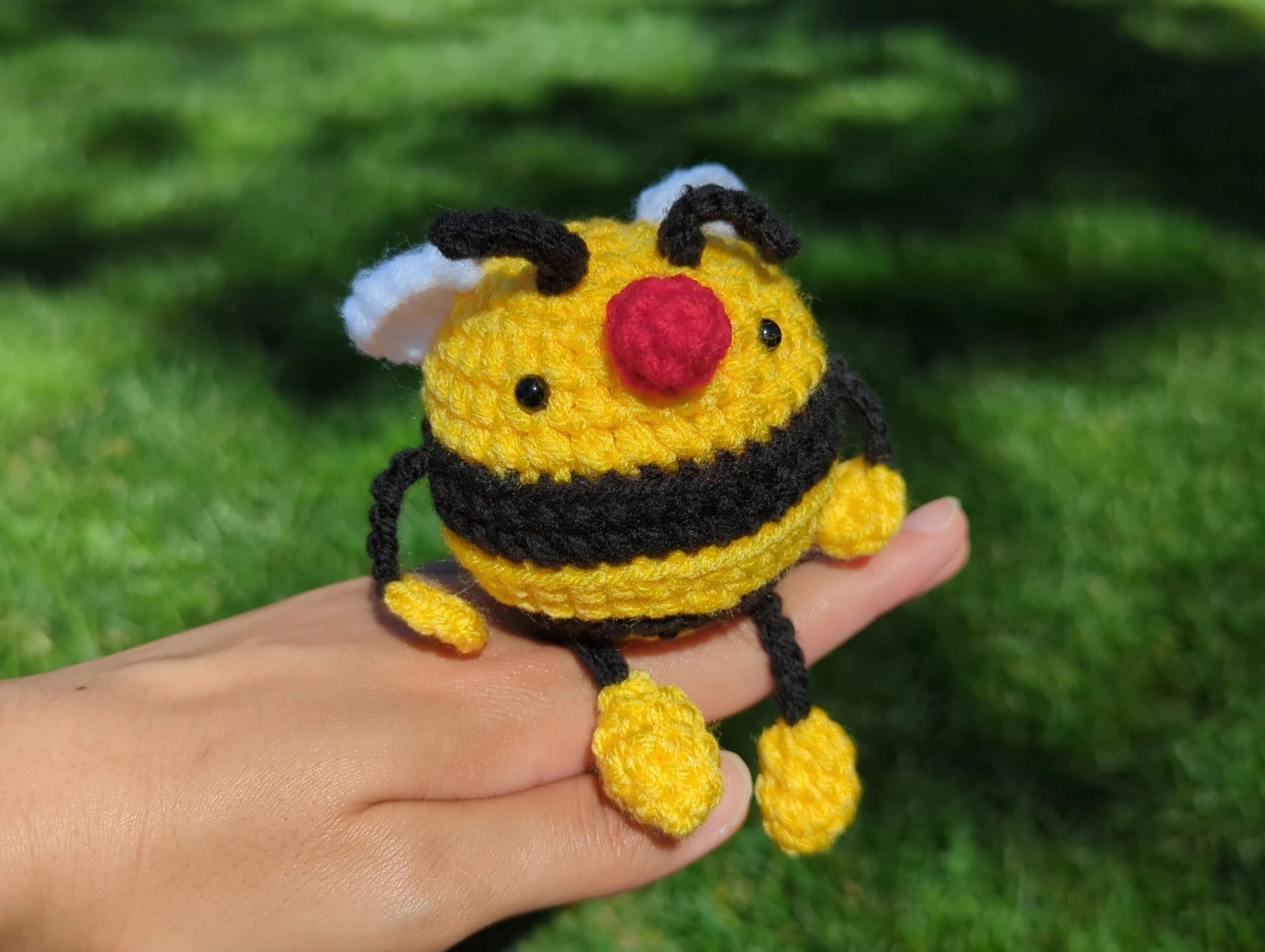 Crocheted soft toy of a cartoon bumble bee sitting on top of a person's hand against a grassy backdrop