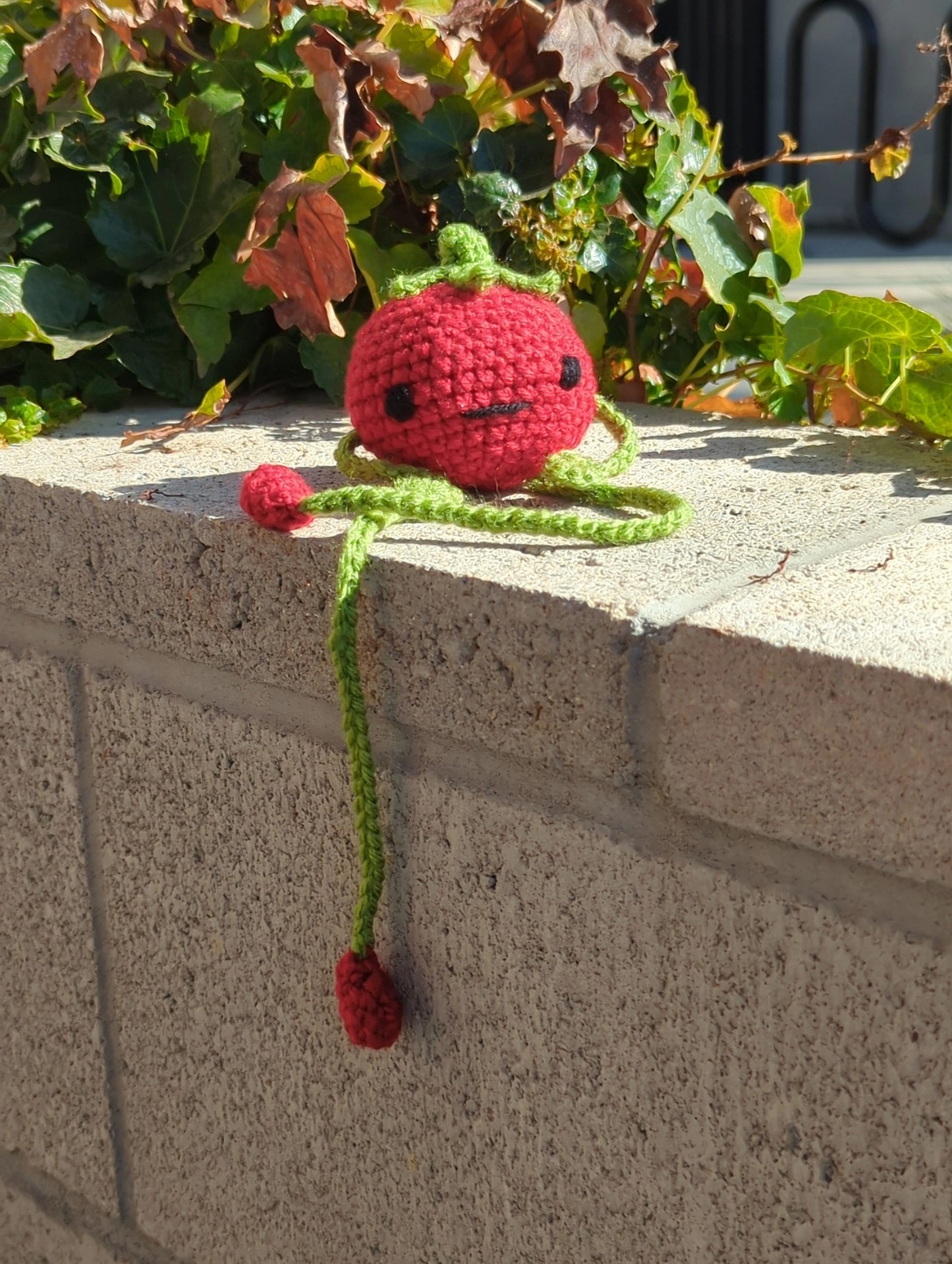 Crocheted soft toy of a cartoon tomato with very long legs sitting on a brick wall covered with ivy