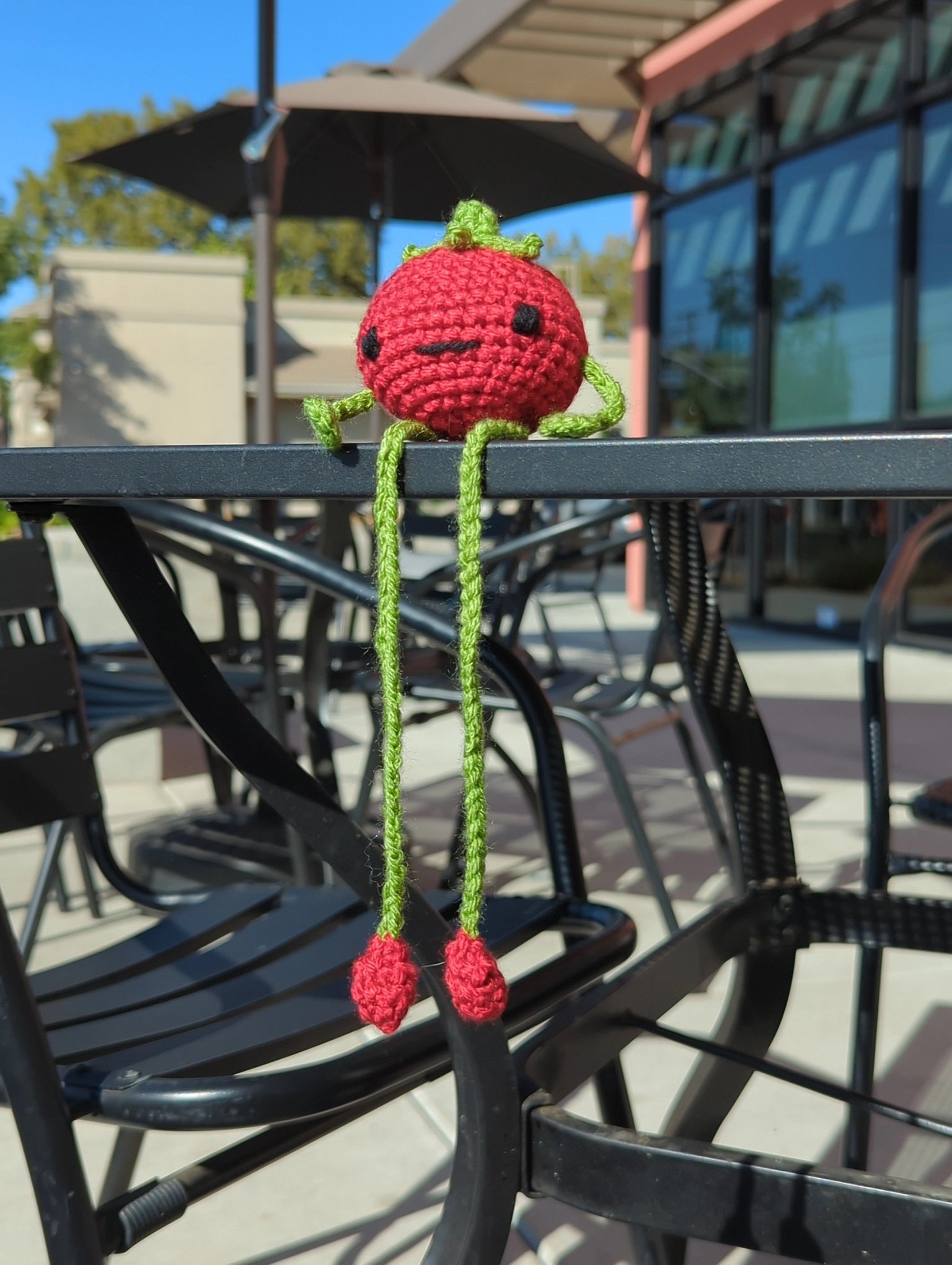 Crocheted soft toy of a cartoon tomato with very long legs sitting on an outdoor cafe table top