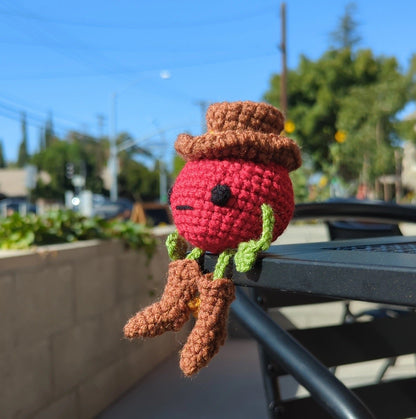 Crocheted soft toy of a cartoon tomato dressed like a cowboy sitting on an outdoor cafe table.