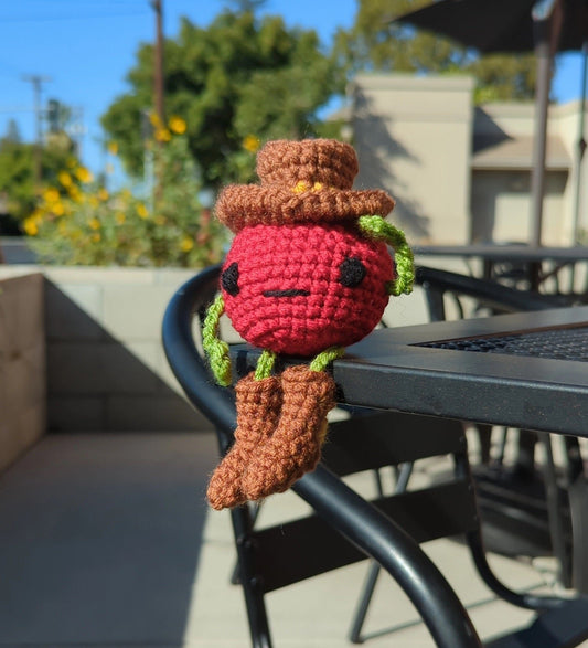 Crocheted soft toy of a cartoon tomato dressed like a cowboy sitting on an outdoor cafe table.