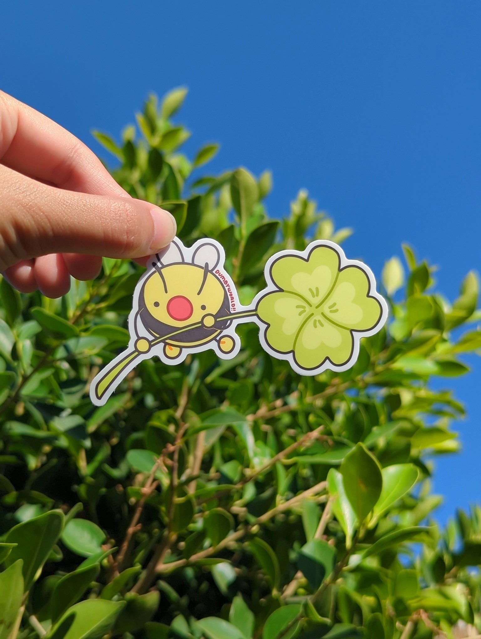 a person's hand holding up a sticker of a cartoon bumble bee holding a clover, held up against a sunny backdrop