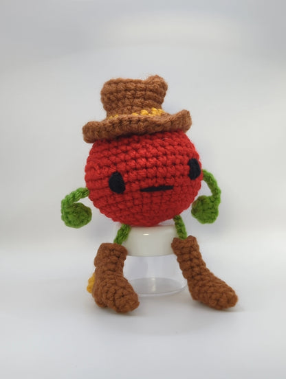 Crocheted soft toy of a cartoon tomato dressed like a cowboy against a white background