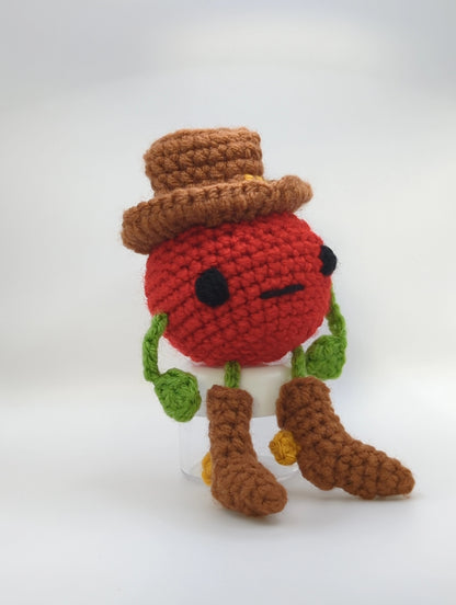 Crocheted soft toy of a cartoon tomato dressed like a cowboy against a white background