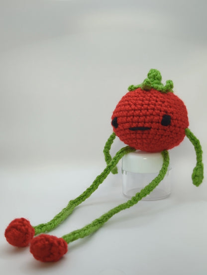 Crocheted soft toy of a cartoon tomato with very long legs sitting against a white backdrop