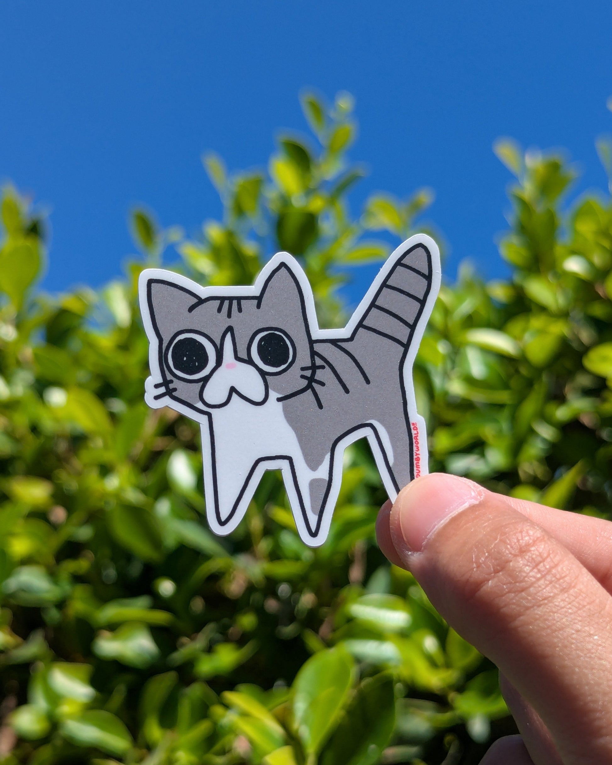Hand holding a cat-shaped sticker with a blue sky and green foliage background