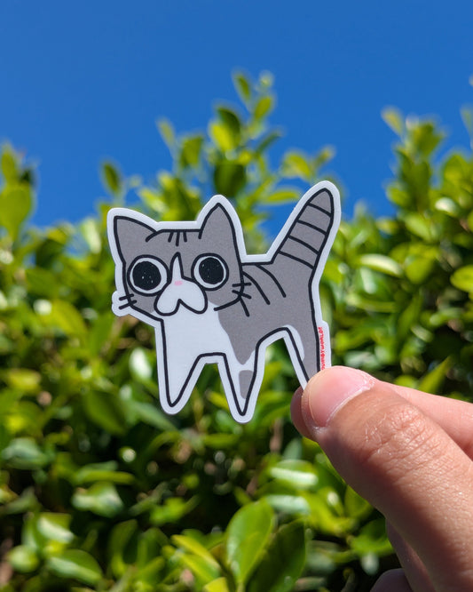 Hand holding a cat-shaped sticker with a blue sky and green foliage background