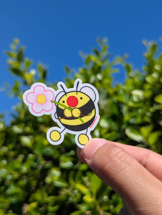 Hand holding a cartoon bee sticker with a flower against a blue sky and green foliage background
