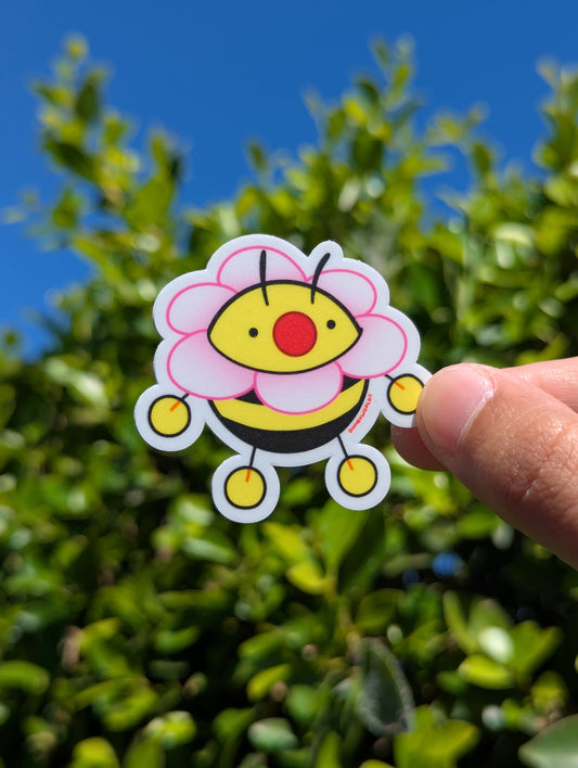 Hand holding a colorful bee-shaped sticker with a floral design against a green leafy background.